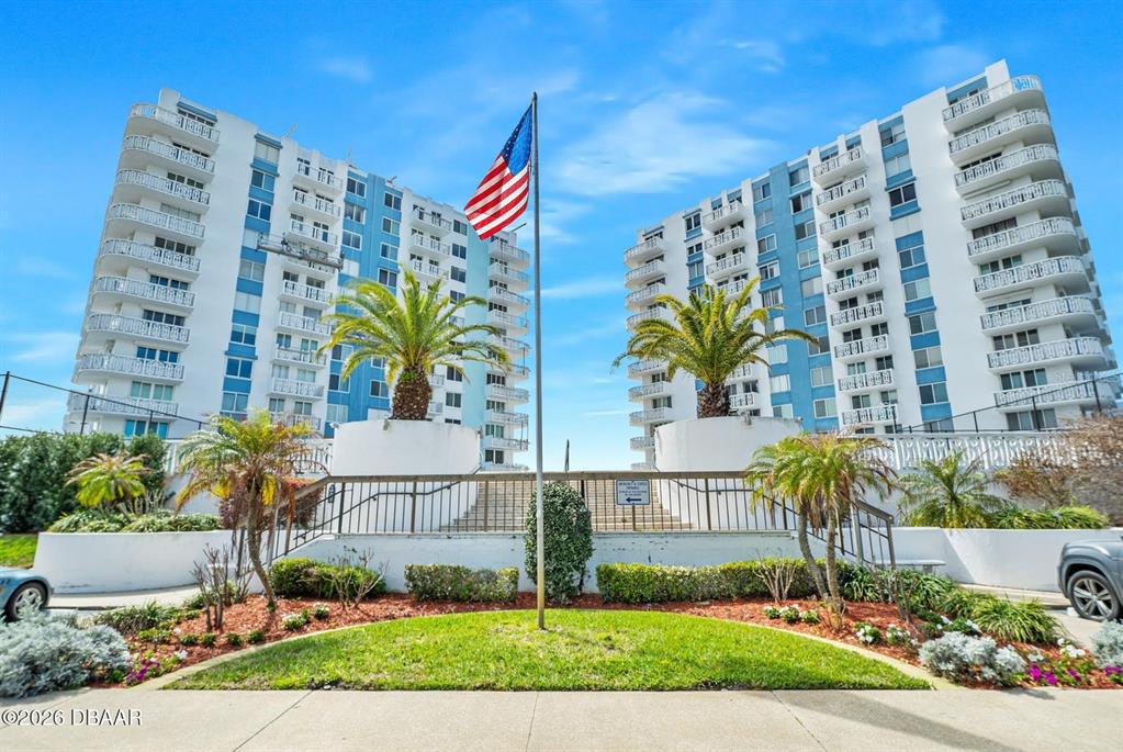 A tall American flag stands between two white multi-story apartment buildings with balconies — 925 N HALIFAX AVENUE UNIT 204, DAYTONA BEACH, FL