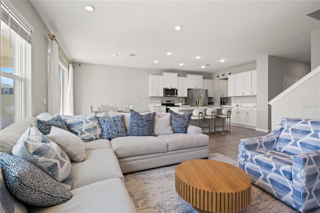 A living room with a sectional sofa and patterned armchair opens to a white kitchen — 6199 WANDERING WILLOW DRIVE, WESLEY CHAPEL, FL
