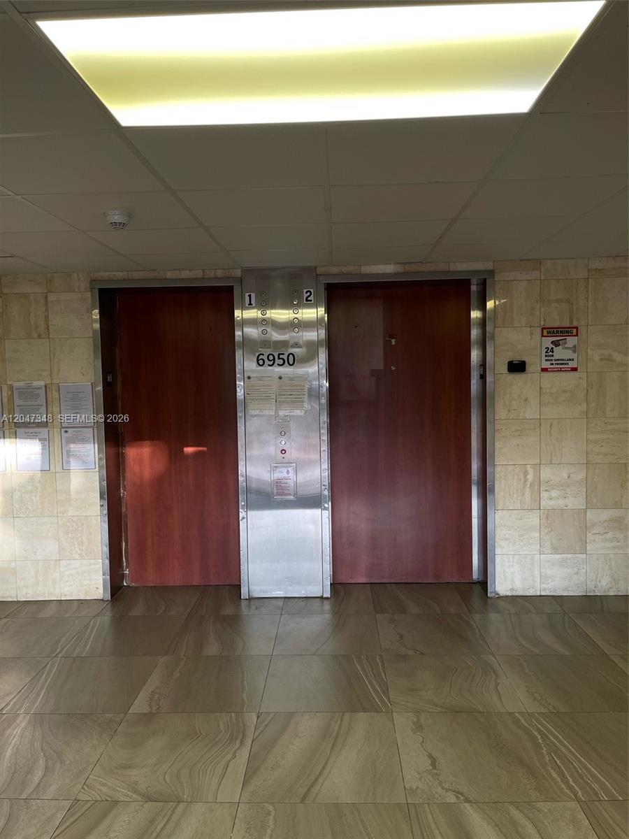 Two elevator doors with wood-grain panels are set into a tiled wall — 6950 Miami Gardens Dr # 312A, Hialeah FL 33015, HIALEAH, FL