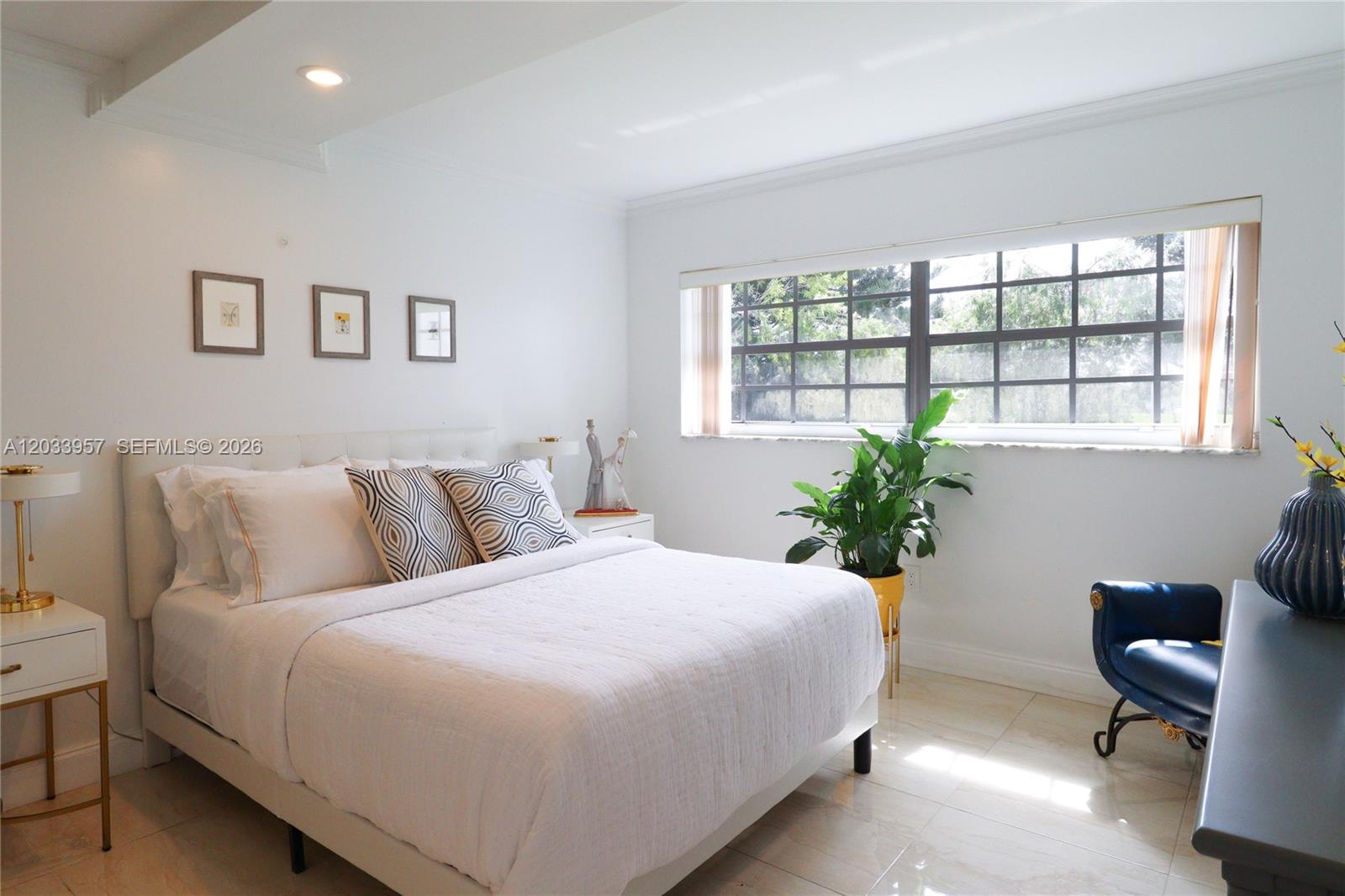 A bright bedroom features a white bed, tiled floors, a large window, and minimalist decor — 7797 Golf Circle Drive # 208J, Margate FL 33063, MARGATE, FL
