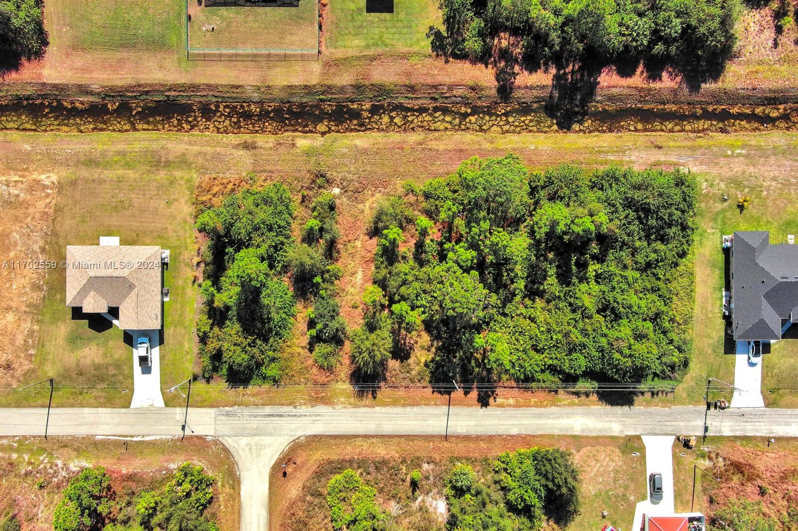 An aerial view shows a wooded vacant lot situated between two residential houses and a canal — 844 Carbon St E, Lehigh Acres FL 33974, LEHIGH ACRES, FL