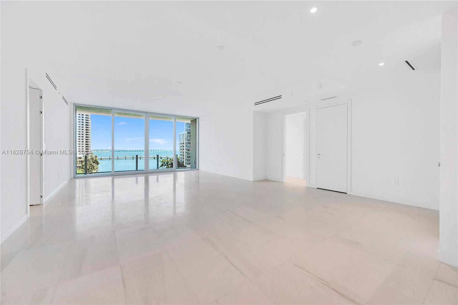 An empty, minimalist living room features white tile floors, recessed lighting, and floor-to-ceiling windows overlooking water — 300 Biscayne Blvd Way # 703, Miami FL 33131, MIAMI, FL