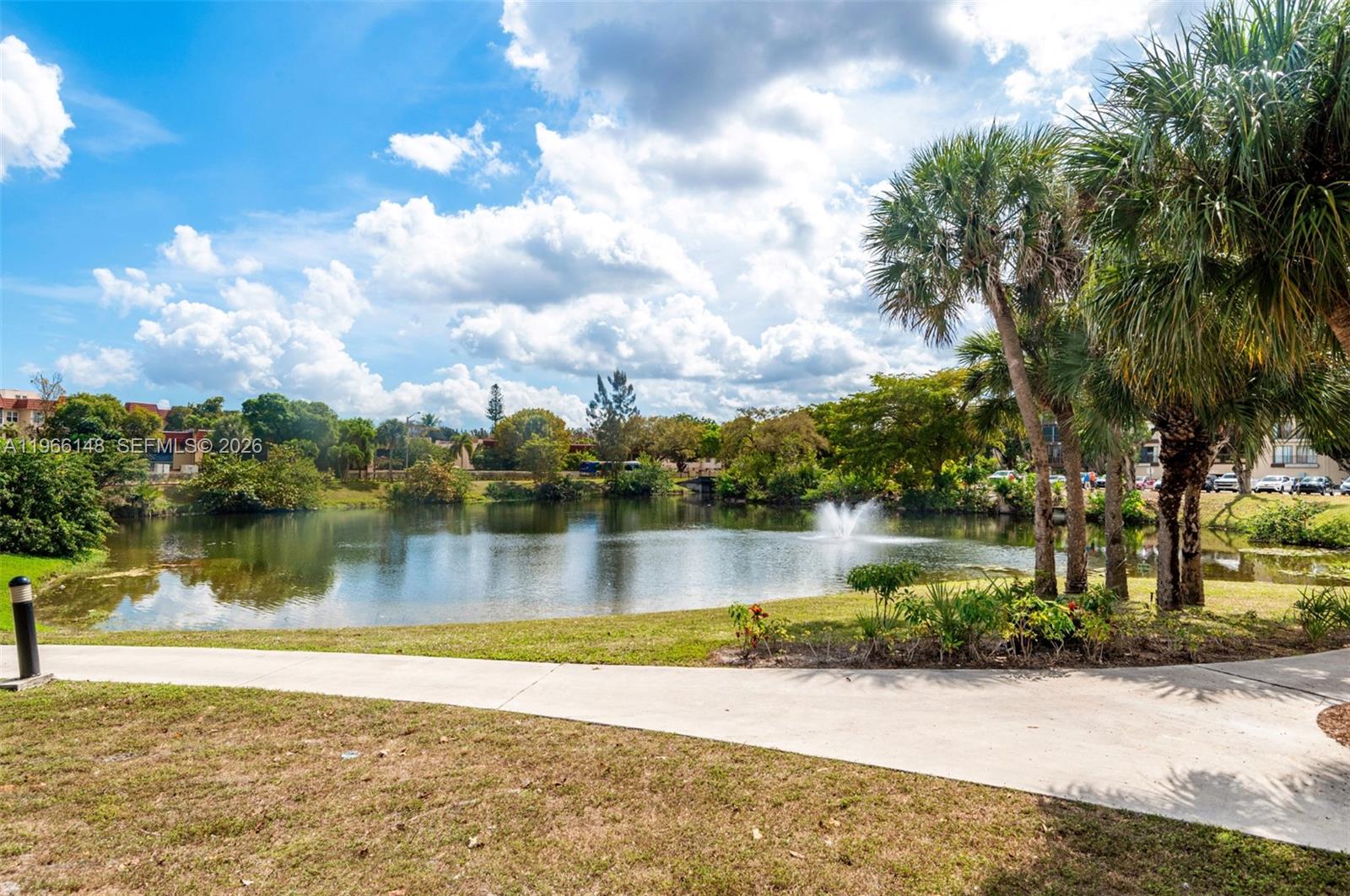 A concrete path leads toward a pond with a fountain surrounded by trees and grass — 6301 N Fall Circle Dr # 213, Lauderhill FL 33319, LAUDERHILL, FL