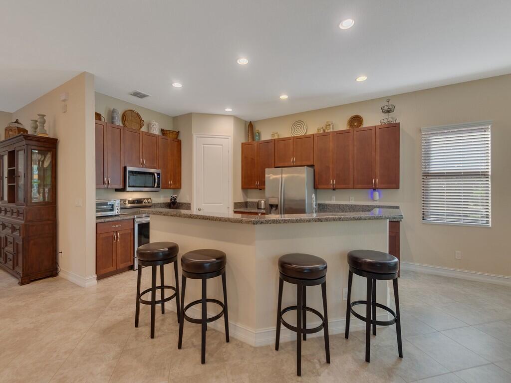 A kitchen features brown cabinets, granite countertops, a breakfast bar with four stools, and tile flooring — 4338 56th Lane, Vero Beach FL 32967, VERO BEACH, FL