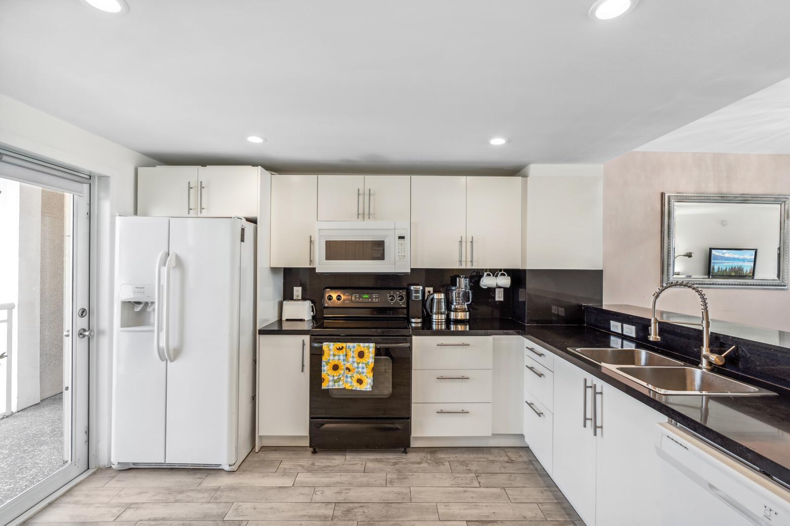 A kitchen features white cabinetry, black countertops, stainless steel appliances, and light wood-look flooring — 750 N Ocean Boulevard # 1507, Pompano Beach FL 33062, POMPANO BEACH, FL
