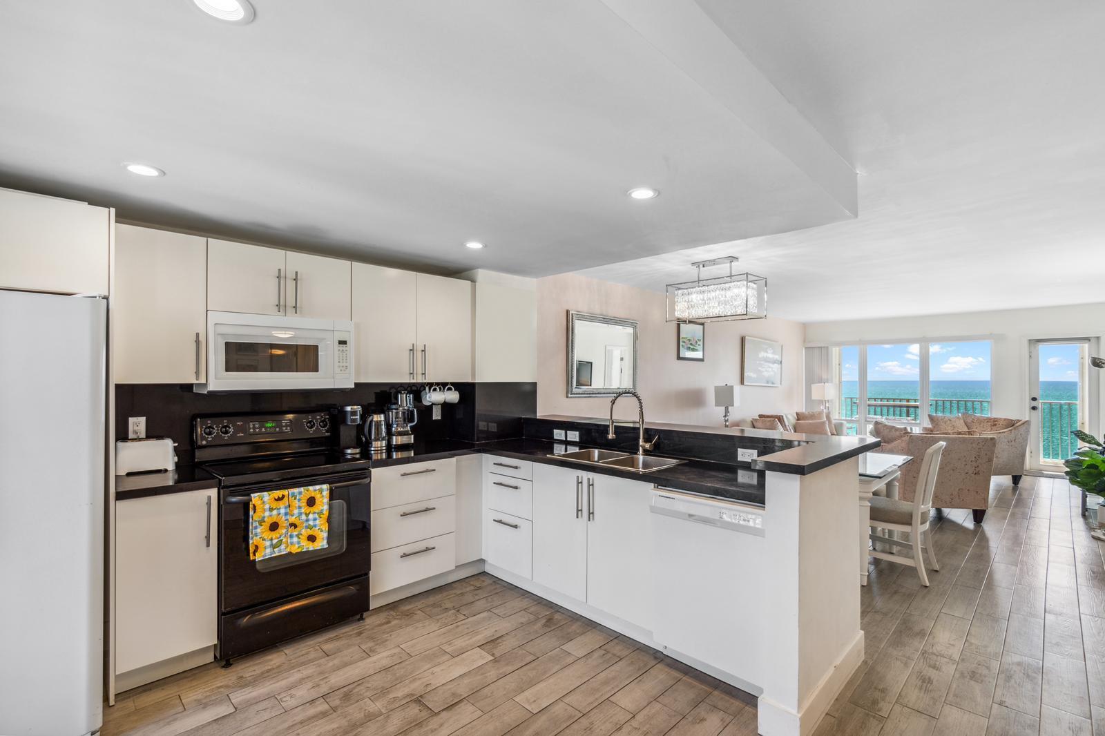 A modern kitchen with white cabinets, black countertops, and stainless steel appliances overlooks a living area — 750 N Ocean Boulevard # 1507, Pompano Beach FL 33062, POMPANO BEACH, FL