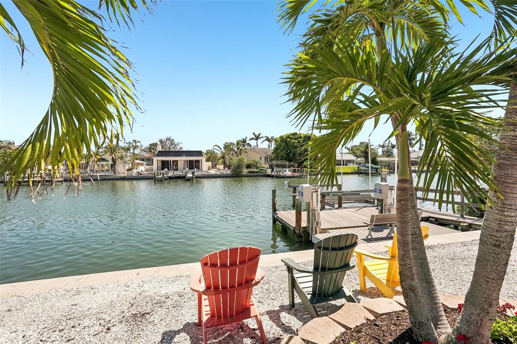 Three colorful Adirondack chairs sit on a gravel shore overlooking a canal with a dock — 85 S POLK DRIVE, SARASOTA, FL