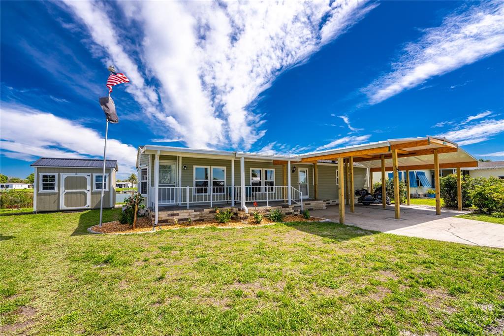 A single-story manufactured home with a covered porch, carport, and detached shed on grass — 6115 SHEARWATER DRIVE, ENGLEWOOD, FL
