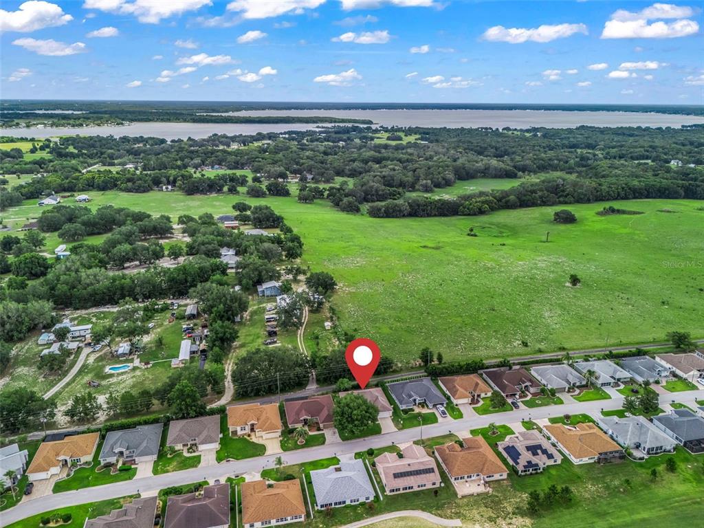 An aerial view shows a suburban neighborhood bordering a large, open green field and lake — 17076 SE 115TH TERRACE ROAD, SUMMERFIELD, FL