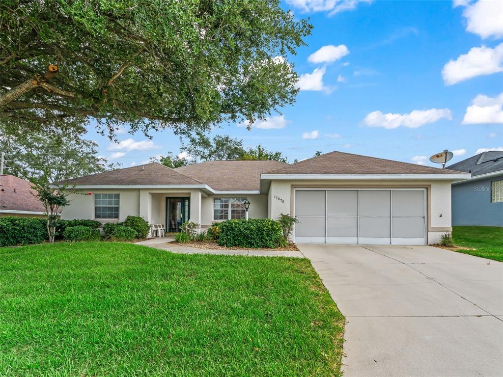 A single-story house with a beige stucco exterior, a two-car garage, and a large tree — 17076 SE 115TH TERRACE ROAD, SUMMERFIELD, FL