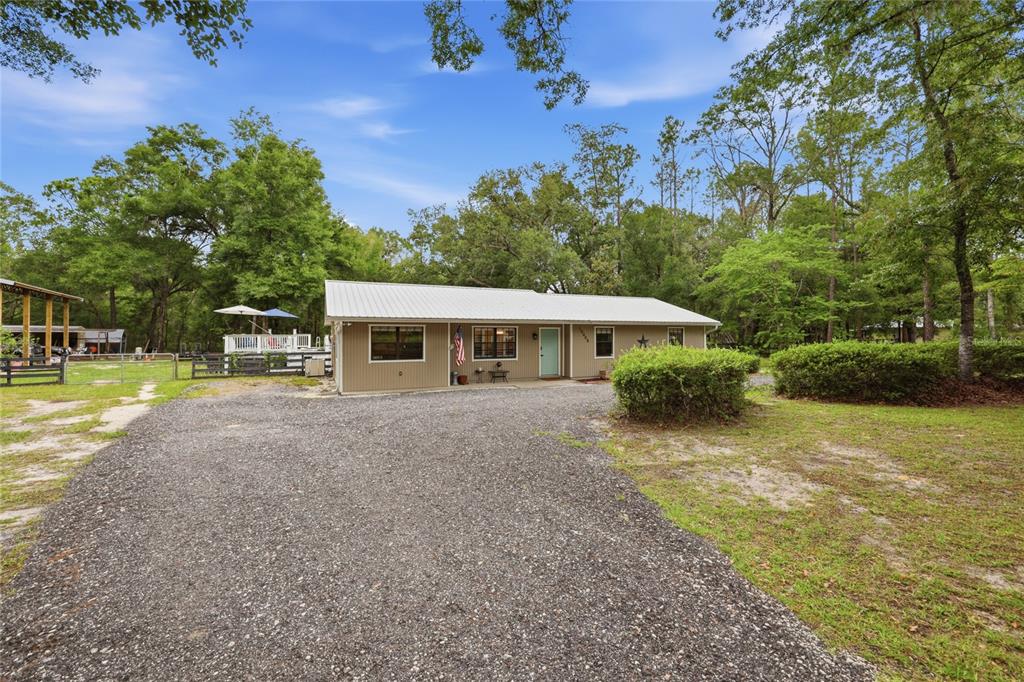 A single-story house with tan siding and a metal roof sits behind a gravel driveway — 10008 SW 80TH WAY, GAINESVILLE, FL