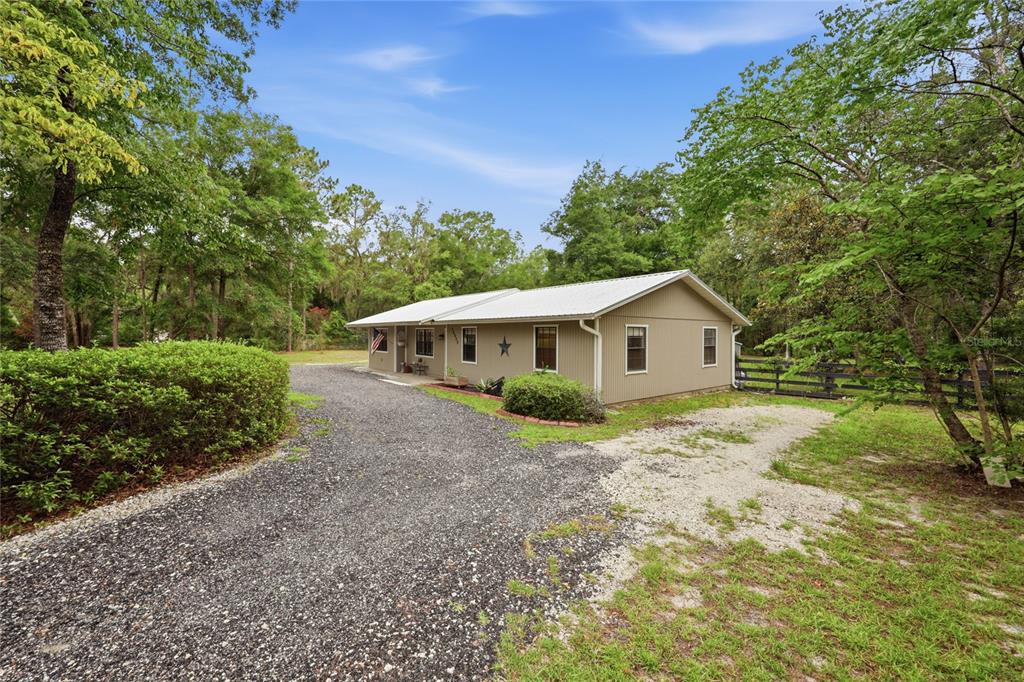 A single-story house with tan siding and a metal roof sits beside a gravel driveway — 10008 SW 80TH WAY, GAINESVILLE, FL