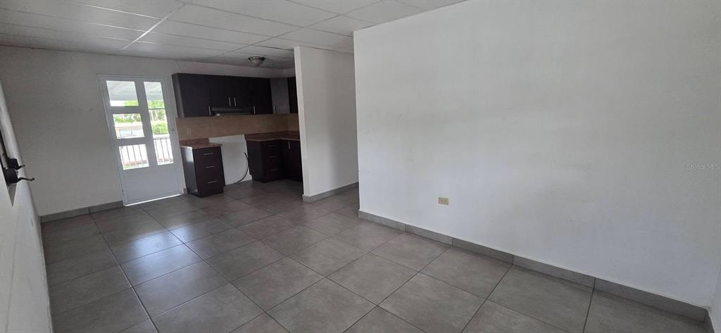A room with gray tile flooring, white walls, and an open kitchen with dark cabinets — Calle 15 C-3 SAN FERNANDO, BAYAMON, PR