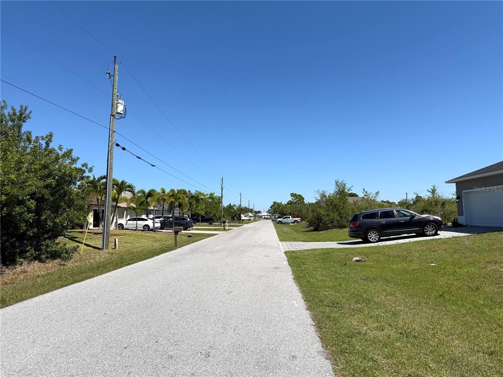 A paved residential street stretches between houses with manicured lawns under a clear blue sky — 9180 SPRING CIRCLE, PORT CHARLOTTE, FL