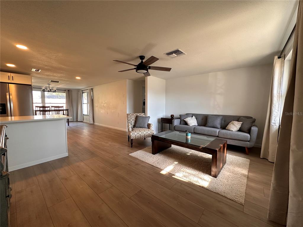 A living room with light wood floors, a grey sofa, and an open kitchen area — 7521 BOUGENVILLE DRIVE, PORT RICHEY, FL