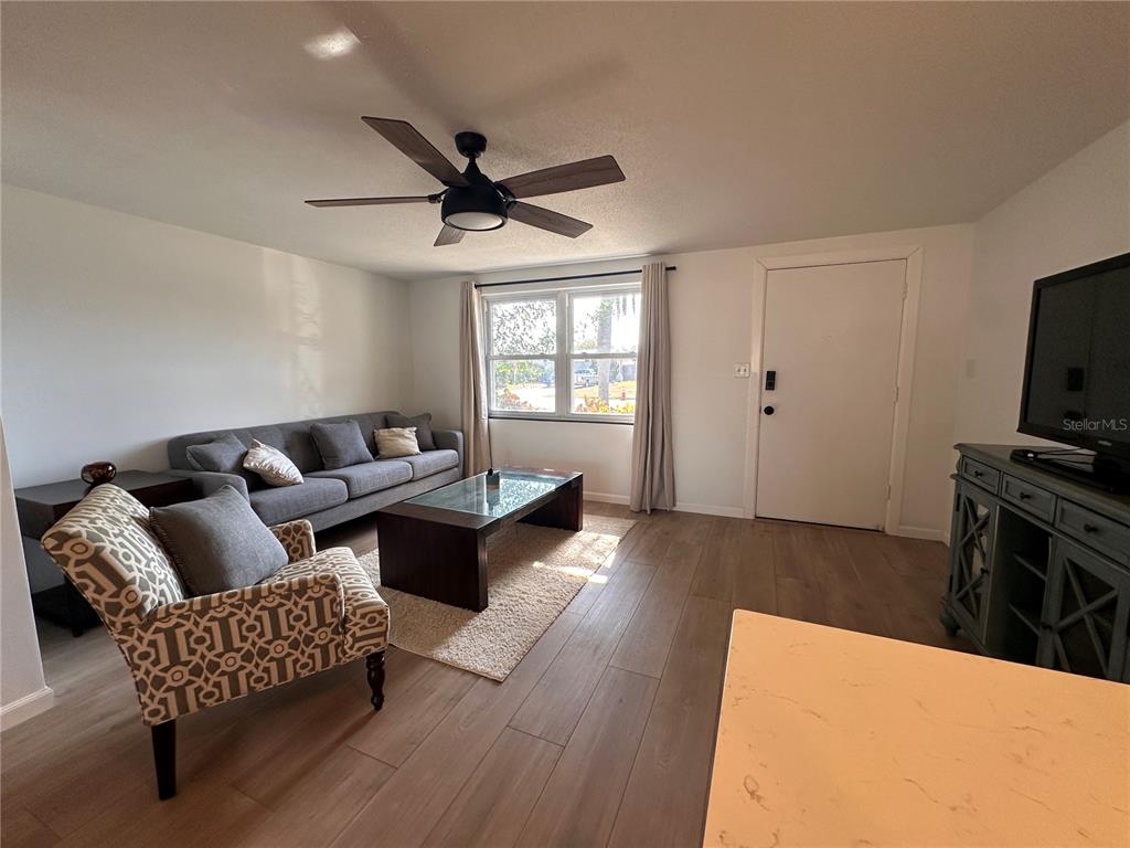 A living room features wood-look flooring, a gray sofa, a patterned armchair, and a ceiling fan — 7521 BOUGENVILLE DRIVE, PORT RICHEY, FL