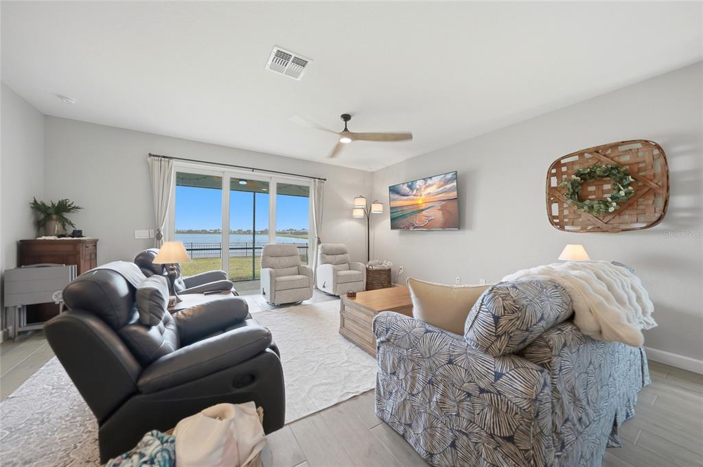 A living room features a patterned sofa, leather recliners, and sliding glass doors overlooking a lake — 8268 SHORE LAKE DRIVE, ENGLEWOOD, FL