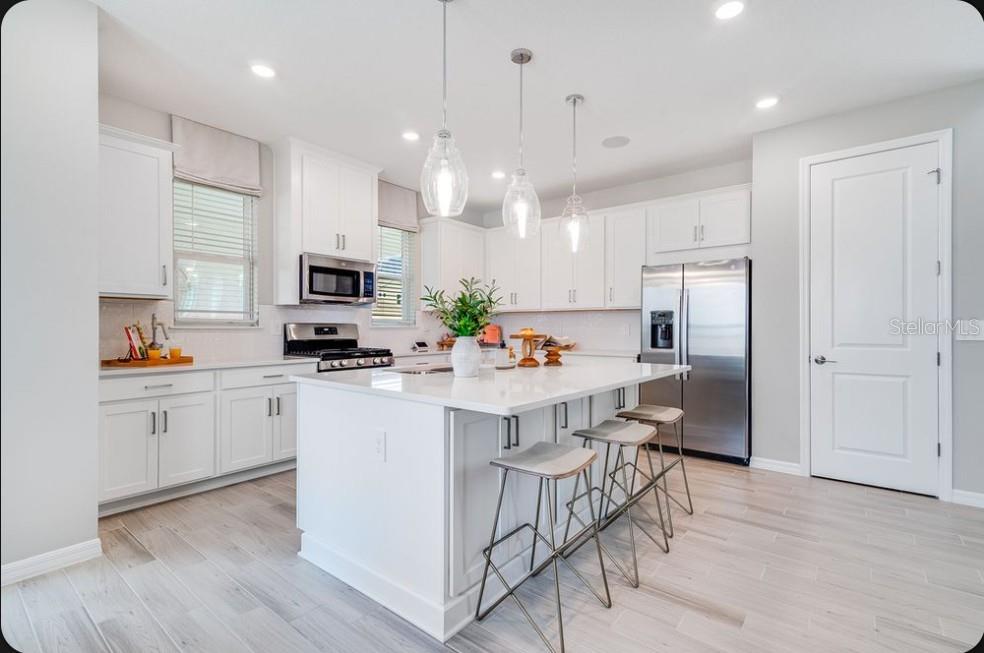 A bright kitchen features white cabinets, a large quartz island, stainless steel appliances, and wood-look flooring — 5180 DOMINION ALLEY, ST CLOUD, FL