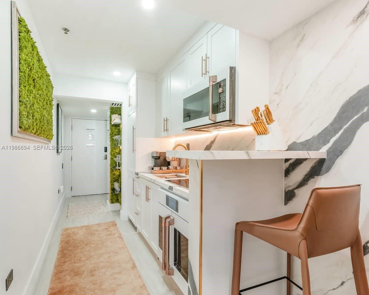 A modern galley kitchen features white cabinetry, marble-style countertops, and a leather bar stool — 100 Collins Ave # 202, Miami Beach FL 33139, MIAMI BEACH, FL
