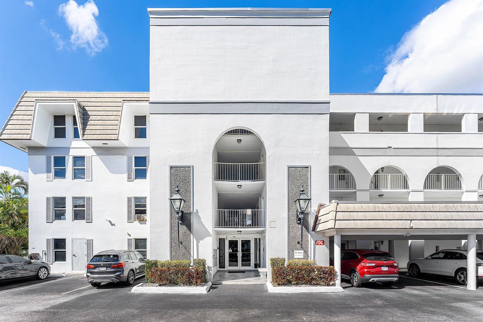 The exterior of a multi-story white apartment building features a central arched entrance and parking — 800 E Camino Real # 409, Boca Raton FL 33432, BOCA RATON, FL