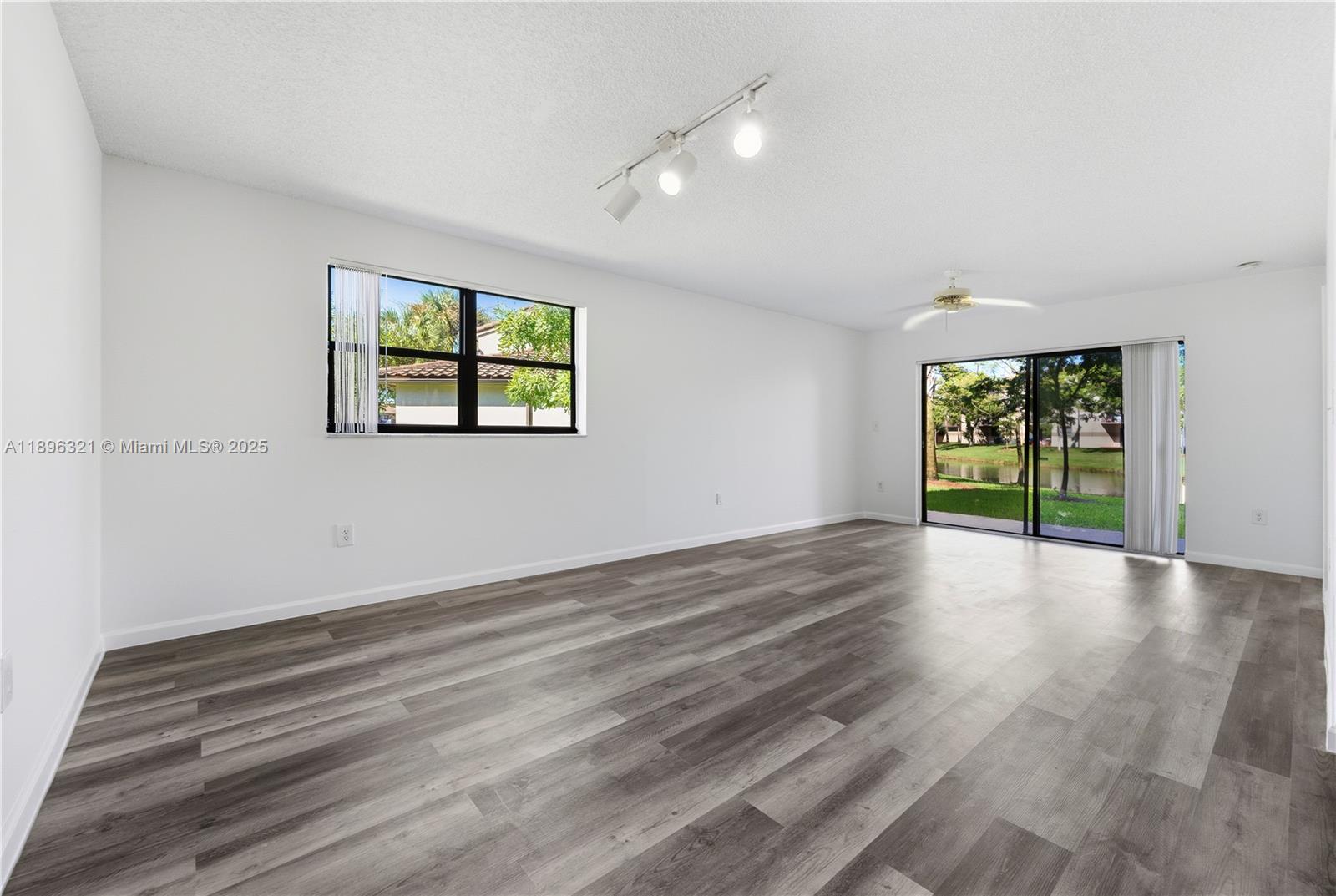An empty living room features gray wood-look flooring, white walls, track lighting, and sliding glass doors — 9877 Nob Hill Ct # 9877, Sunrise FL 33351, SUNRISE, FL
