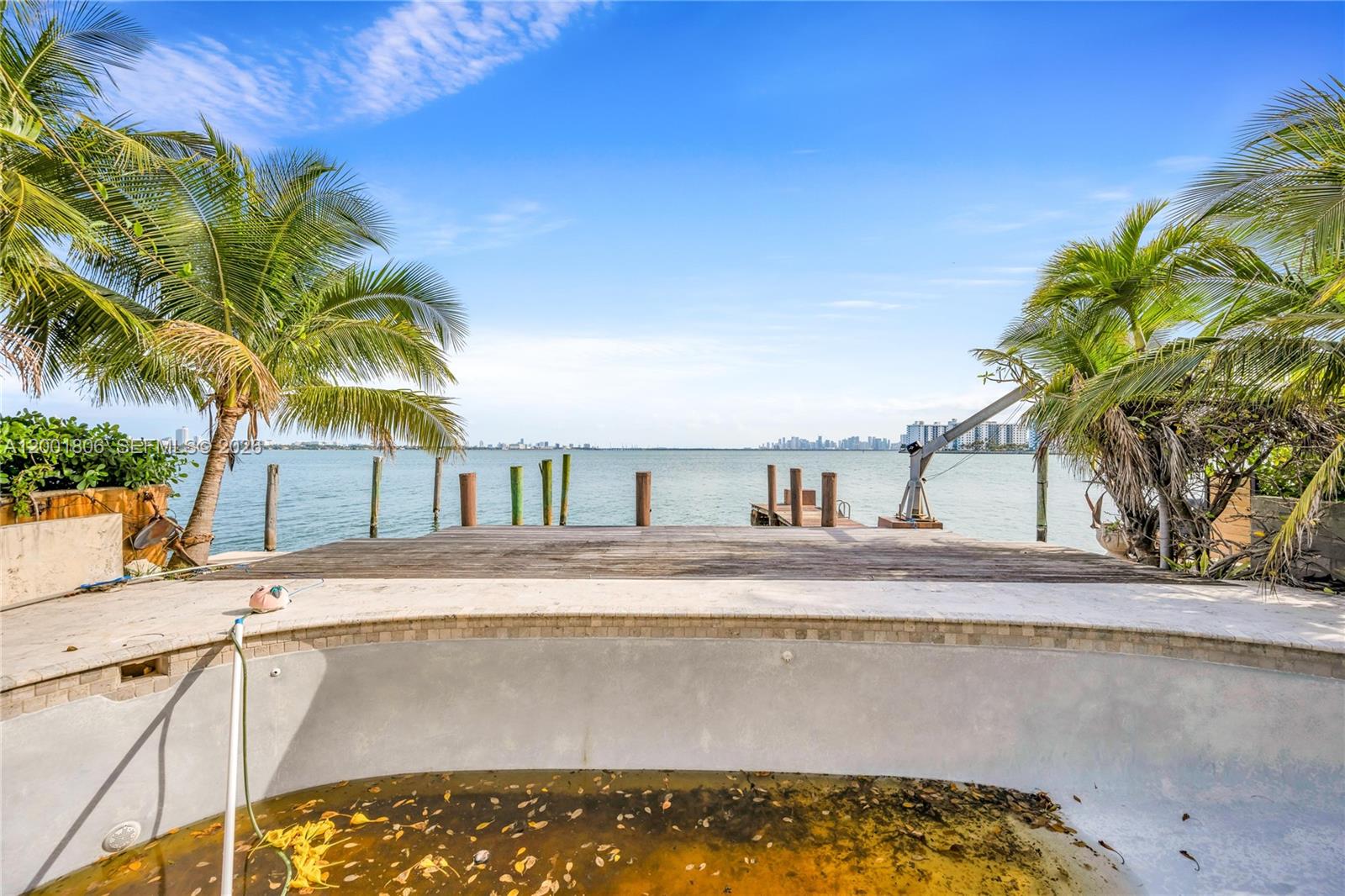 A neglected swimming pool overlooks a wooden deck and a bay with a distant skyline — MIAMI BEACH, FL