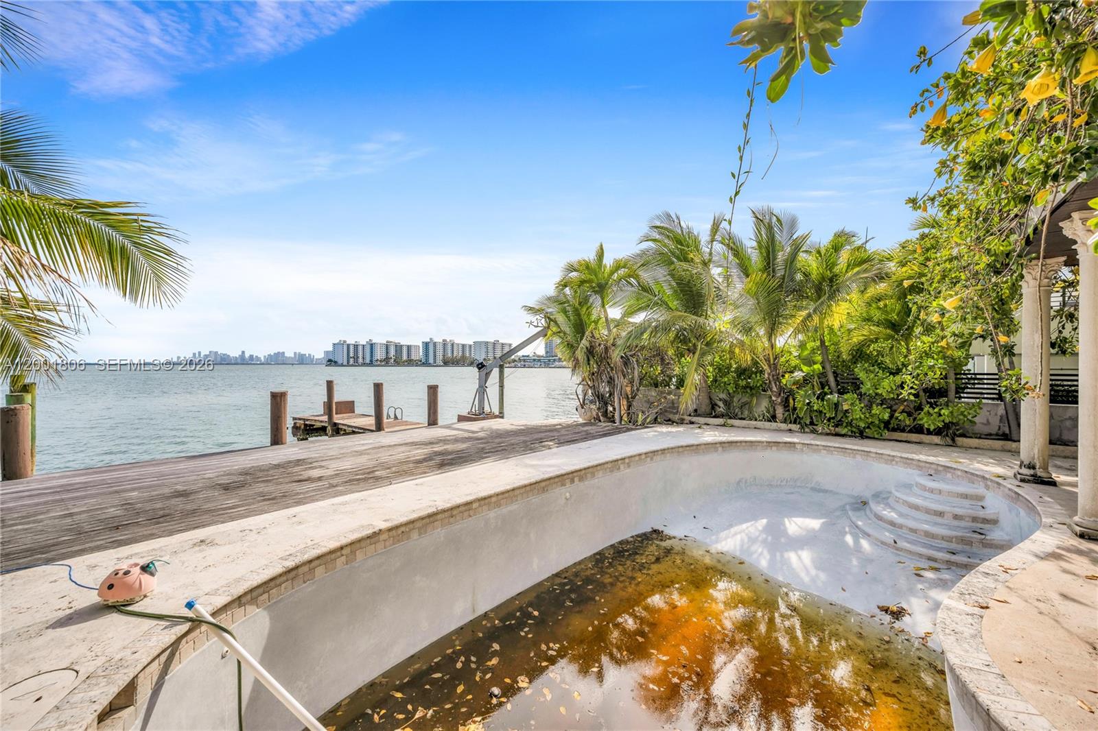 An empty, dirty swimming pool sits on a wooden deck overlooking a bay and skyline — MIAMI BEACH, FL