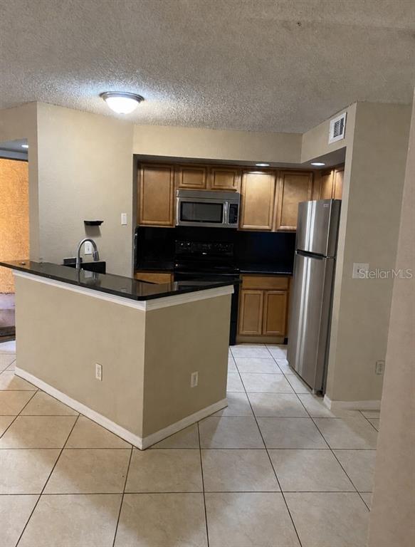A kitchen features light wood cabinets, black countertops, stainless steel appliances, and tile flooring — 4524 COMMANDER DRIVE UNIT 1615, ORLANDO, FL