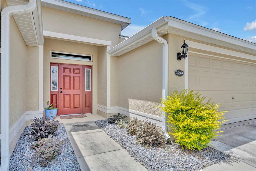 A beige house exterior features a red front door, a garage, and gravel landscaping — 26669 OTTER CREEK LANE, LEESBURG, FL