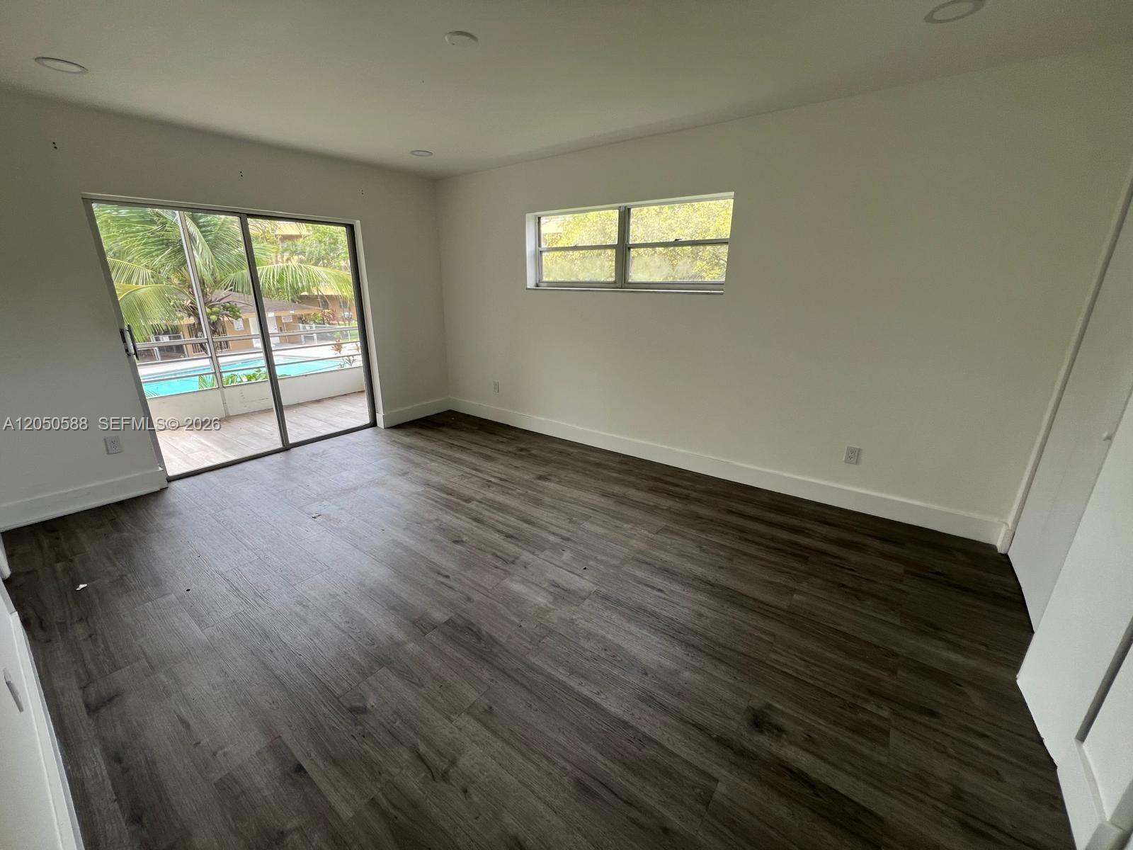 An empty bedroom features dark wood-look flooring, white walls, a sliding glass door, and windows — 4111 Stirling Rd # 209, Dania Beach FL 33314, DANIA BEACH, FL