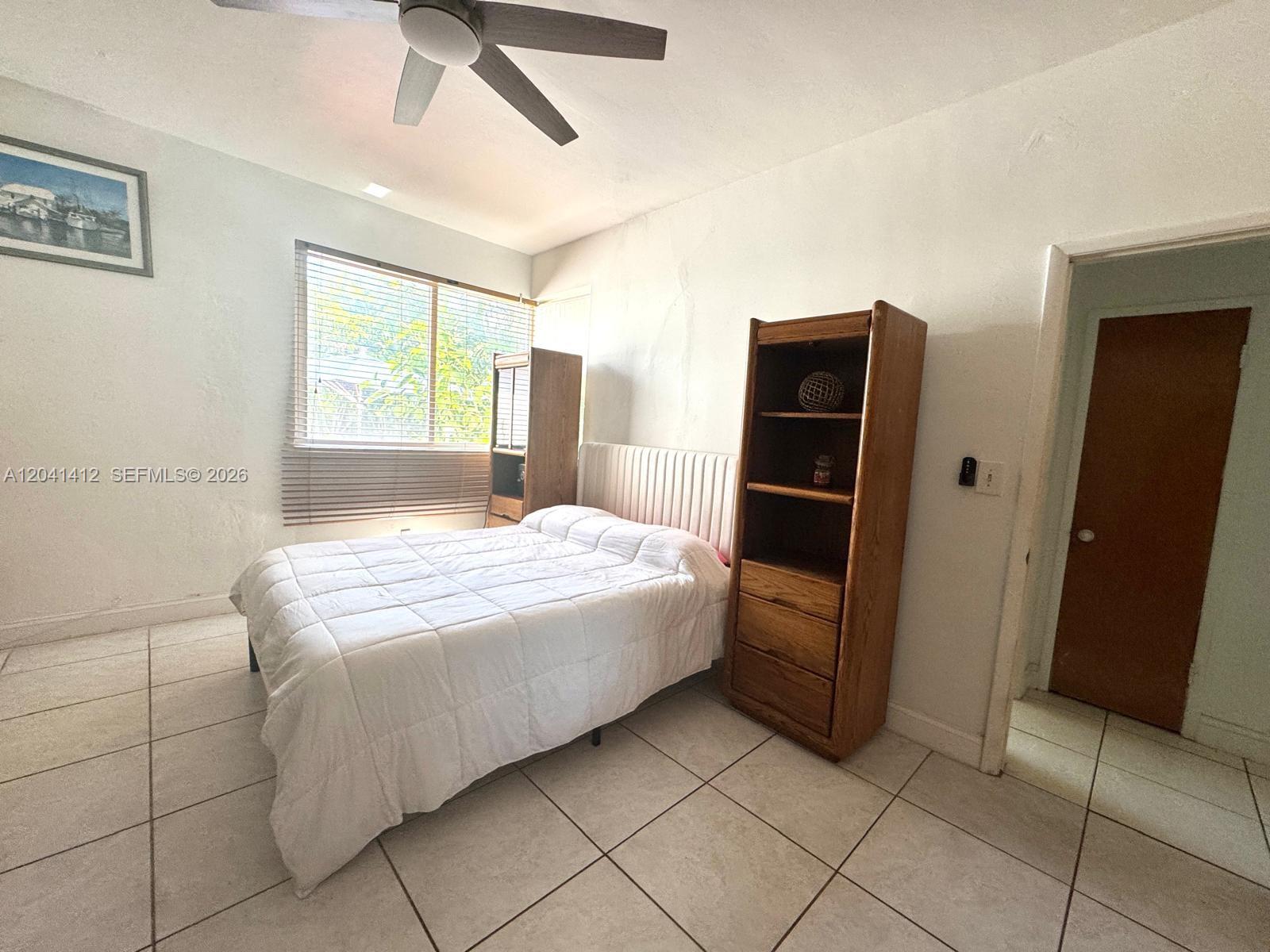 A bedroom features a bed, two wooden shelving units, a ceiling fan, and tiled flooring — BAY HARBOR ISLANDS, FL