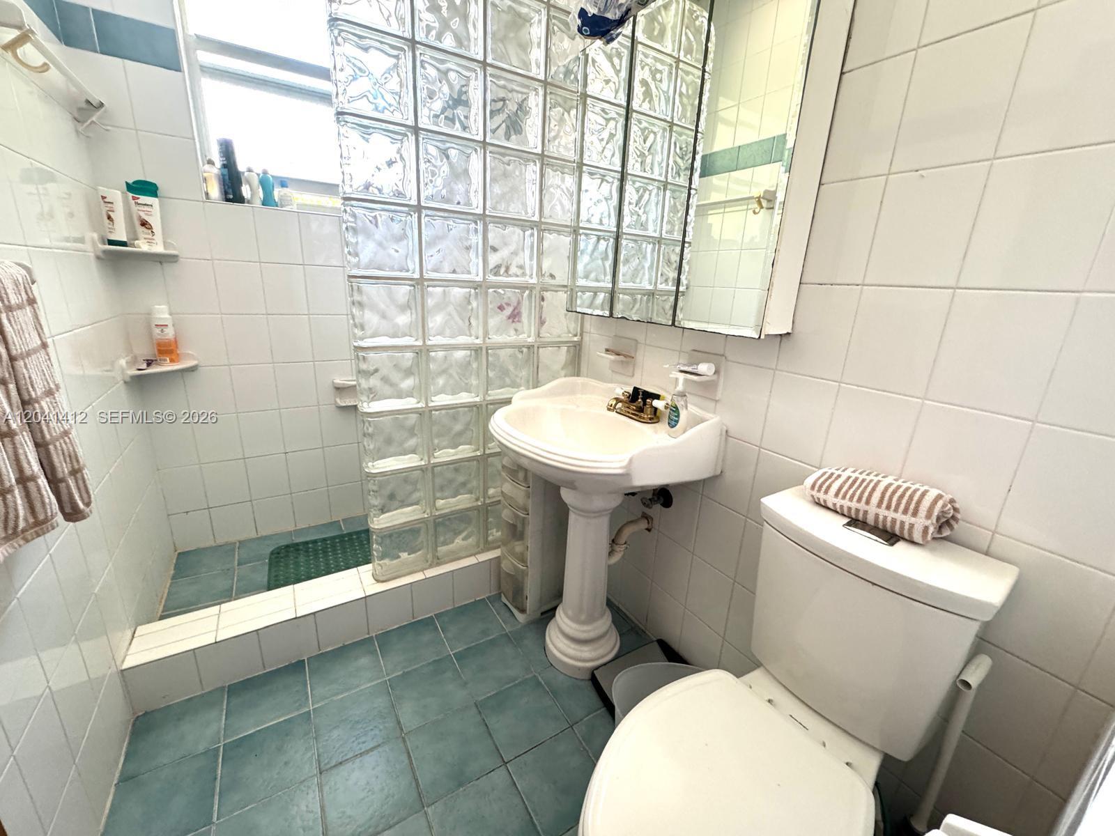A bathroom features a pedestal sink, a glass block shower wall, and white tiled walls — BAY HARBOR ISLANDS, FL