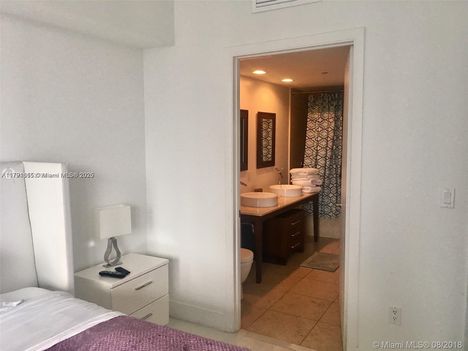 A bedroom with a white nightstand leads into a bathroom featuring dual vessel sinks — 485 Brickell Ave # 1903, Miami FL 33131, MIAMI, FL
