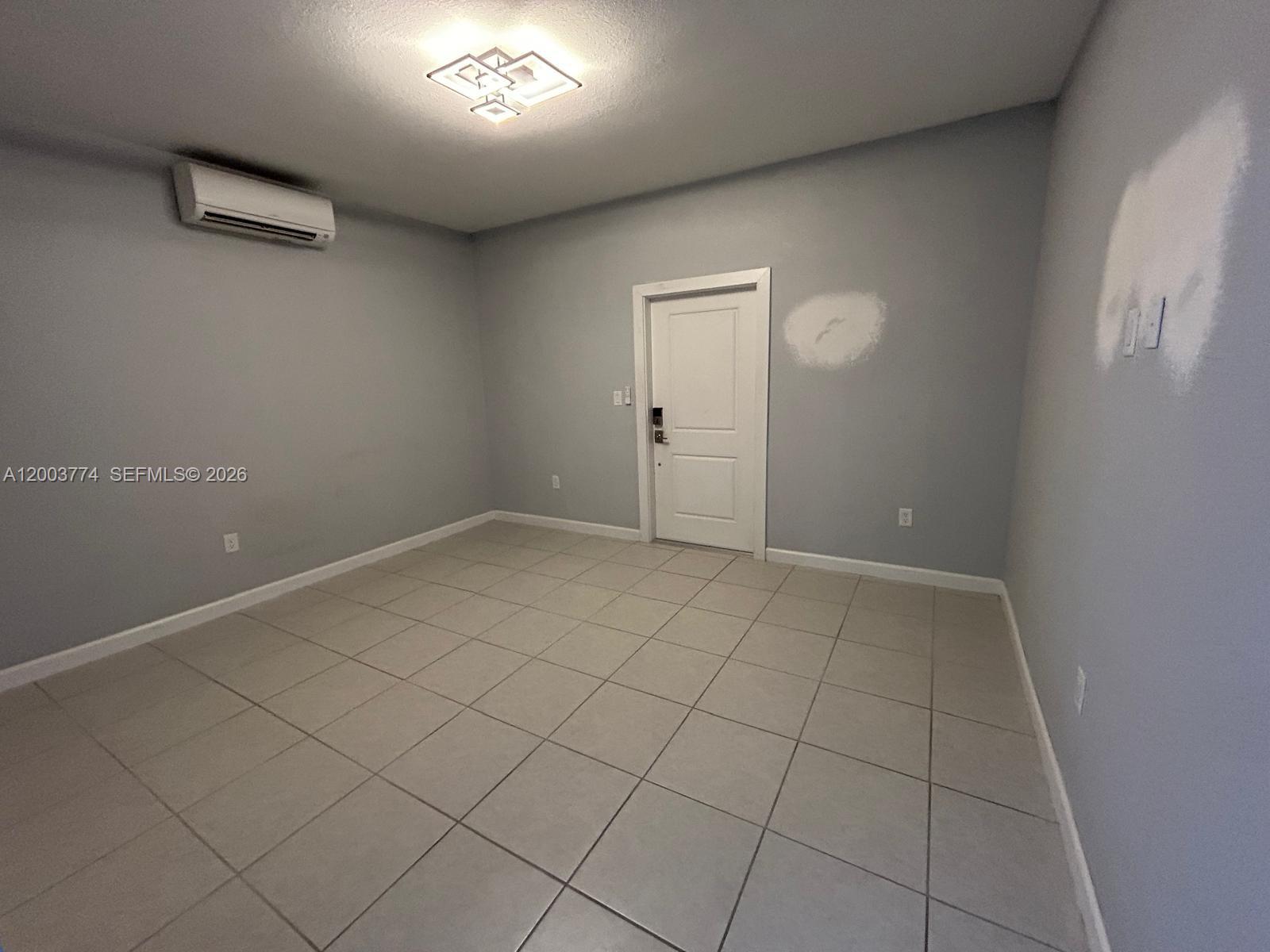 An empty room features gray walls, light tile flooring, a white door, and a mini-split unit — HOMESTEAD, FL