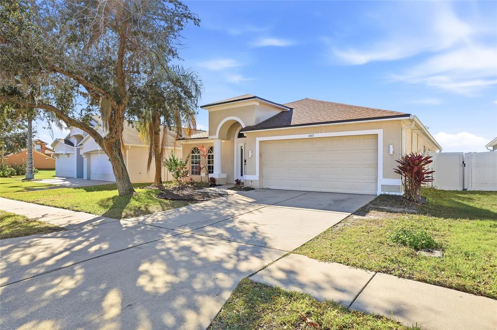 A single-story suburban house with a tan stucco exterior, a two-car garage, and a driveway — 4145 MEDBURY DRIVE, WESLEY CHAPEL, FL