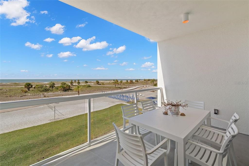 A balcony with white patio furniture overlooks a grassy lawn, sandy beach, and the ocean — 11500 GULF BOULEVARD UNIT 304, TREASURE ISLAND, FL