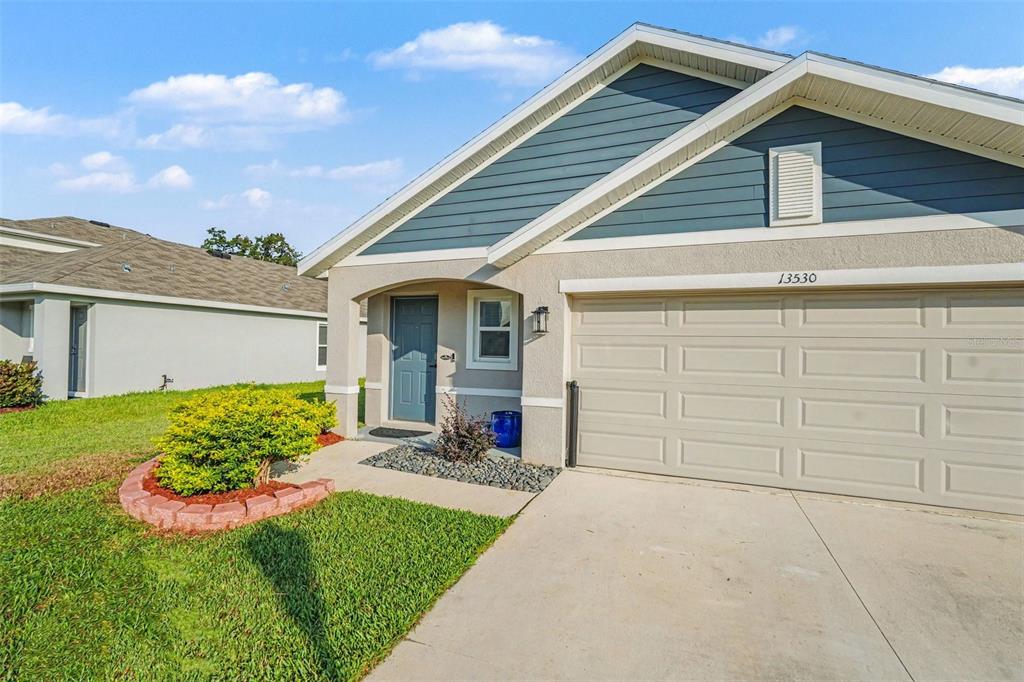 A single-story house exterior features a beige garage, blue front door, and a landscaped yard — 13530 MOUNT IVY COURT, DADE CITY, FL