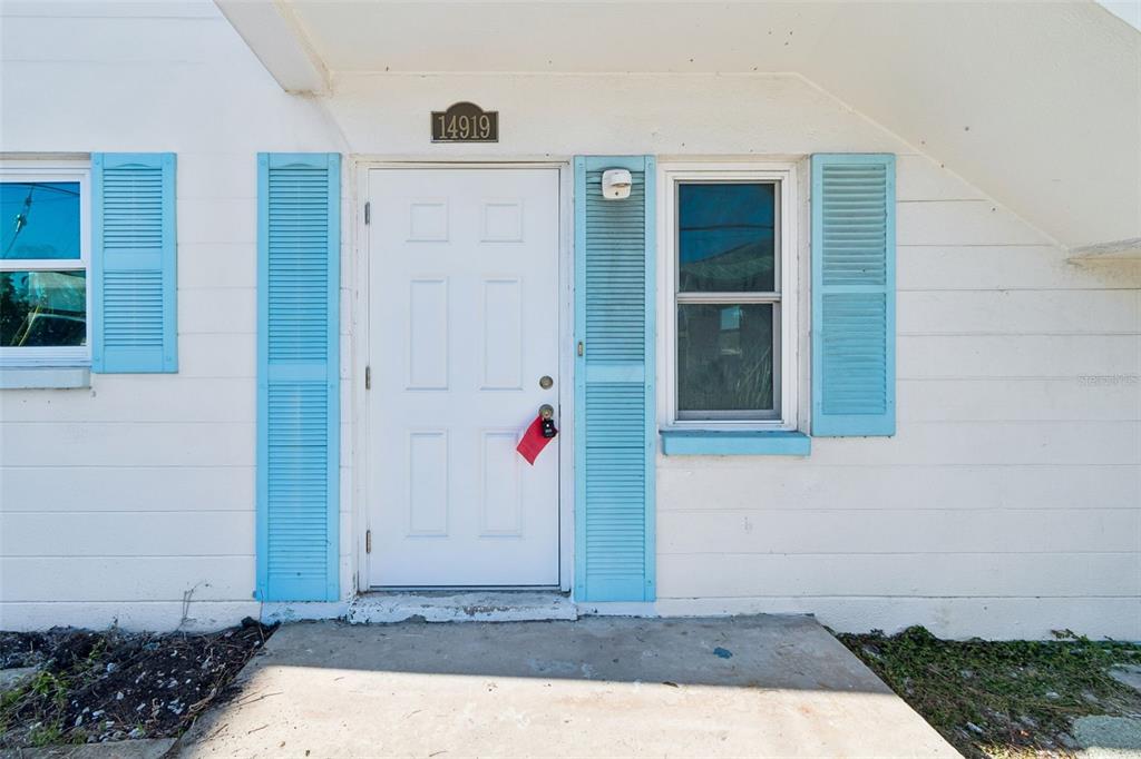 A white exterior wall features a front door with a lockbox and blue shutters — 14919 N BAYSHORE DRIVE UNIT 1, MADEIRA BEACH, FL