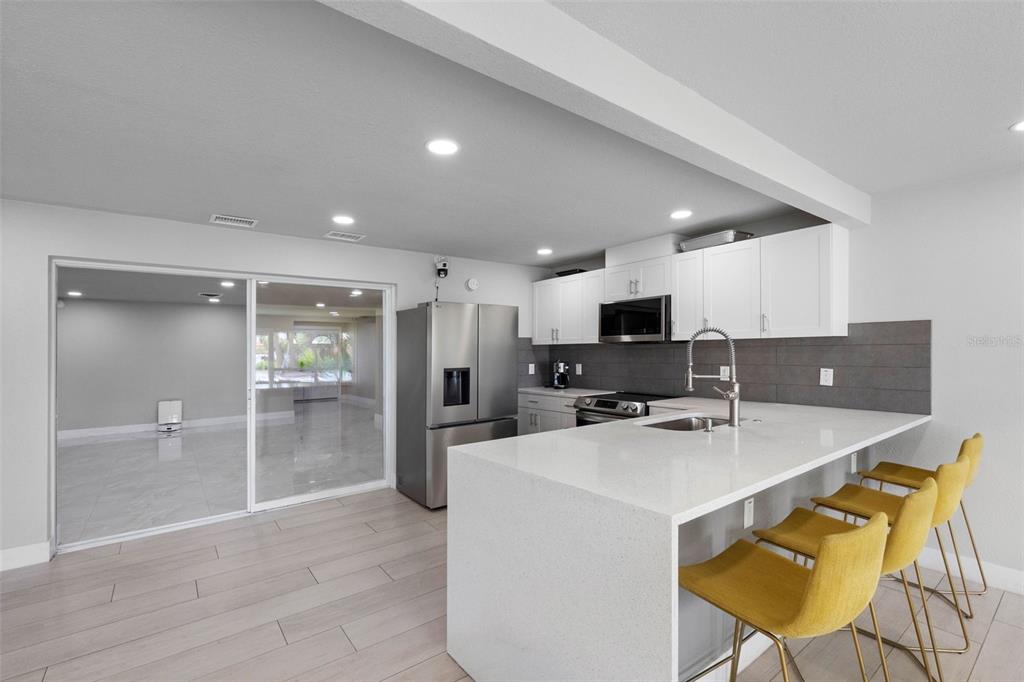 A modern kitchen features white cabinets, a quartz island with yellow stools, and tile flooring — 7032 MORNINGSTAR LANE, NEW PORT RICHEY, FL