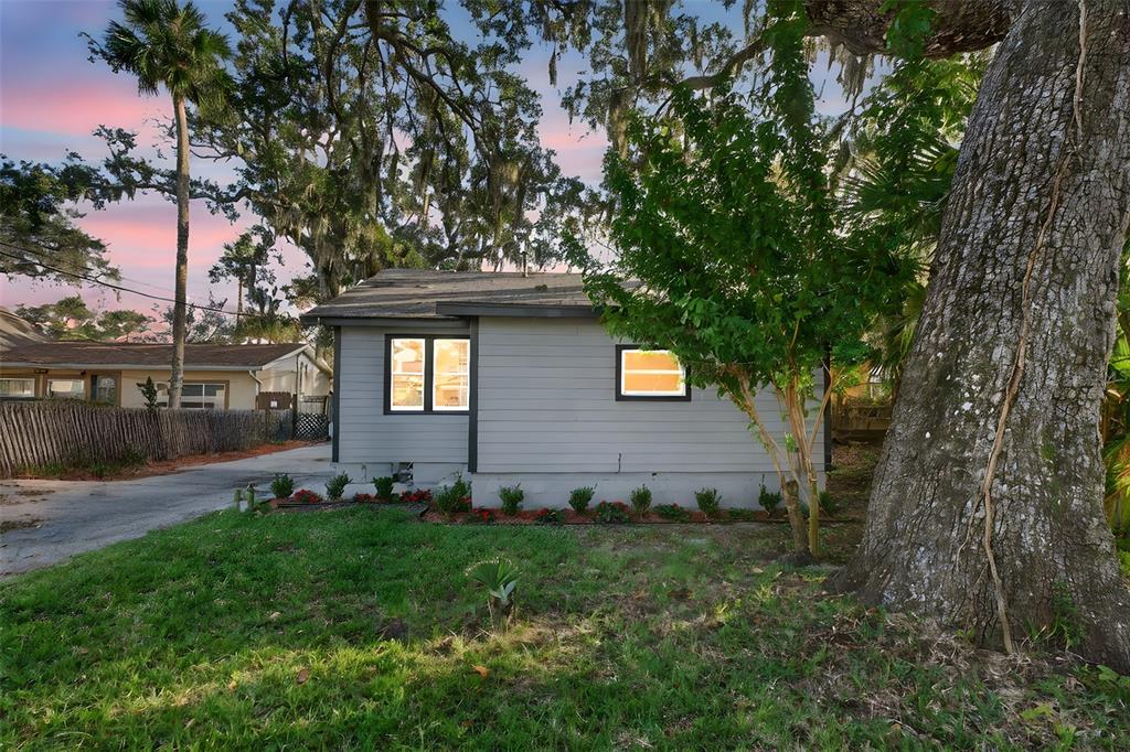 A gray single-story house exterior features a small lawn, mature trees, and a paved driveway — 115 PALM PLACE, DAYTONA BEACH, FL