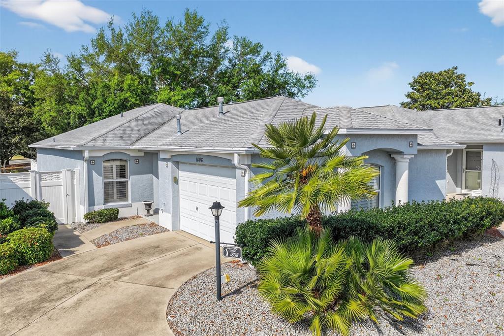 A light blue single-story house features a white garage, a palm tree, and gravel landscaping — 1808 PEACHTREE AVENUE, THE VILLAGES, FL