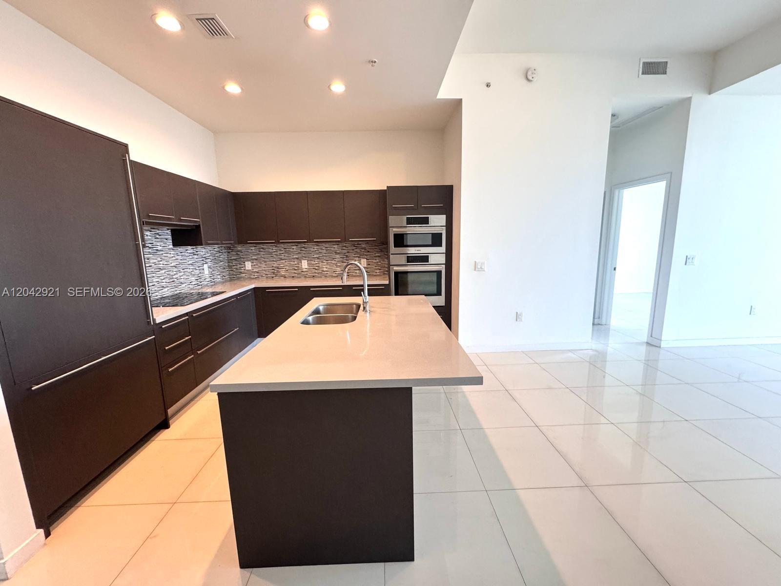 A modern kitchen features dark cabinetry, a central island with a sink, and white flooring — 7825 NW 107th Ave # 706, Doral FL 33178, DORAL, FL