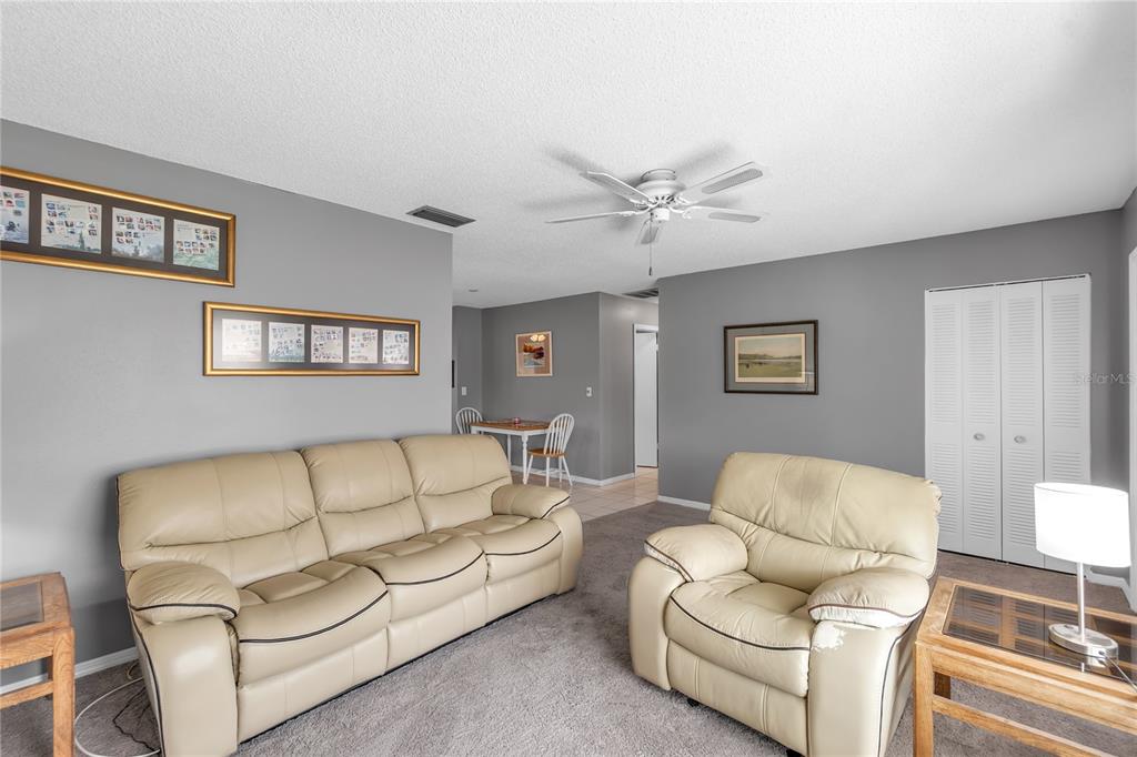A living room features a beige leather sofa and armchair on gray carpeted flooring — 702 HUXLEY PLACE, SUN CITY CENTER, FL