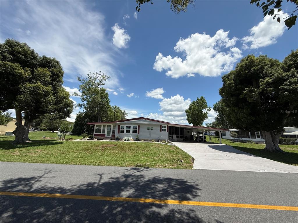 A single-story mobile home with a carport and a large front lawn under a blue sky — 19 MAGNOLIA LANE, WILDWOOD, FL