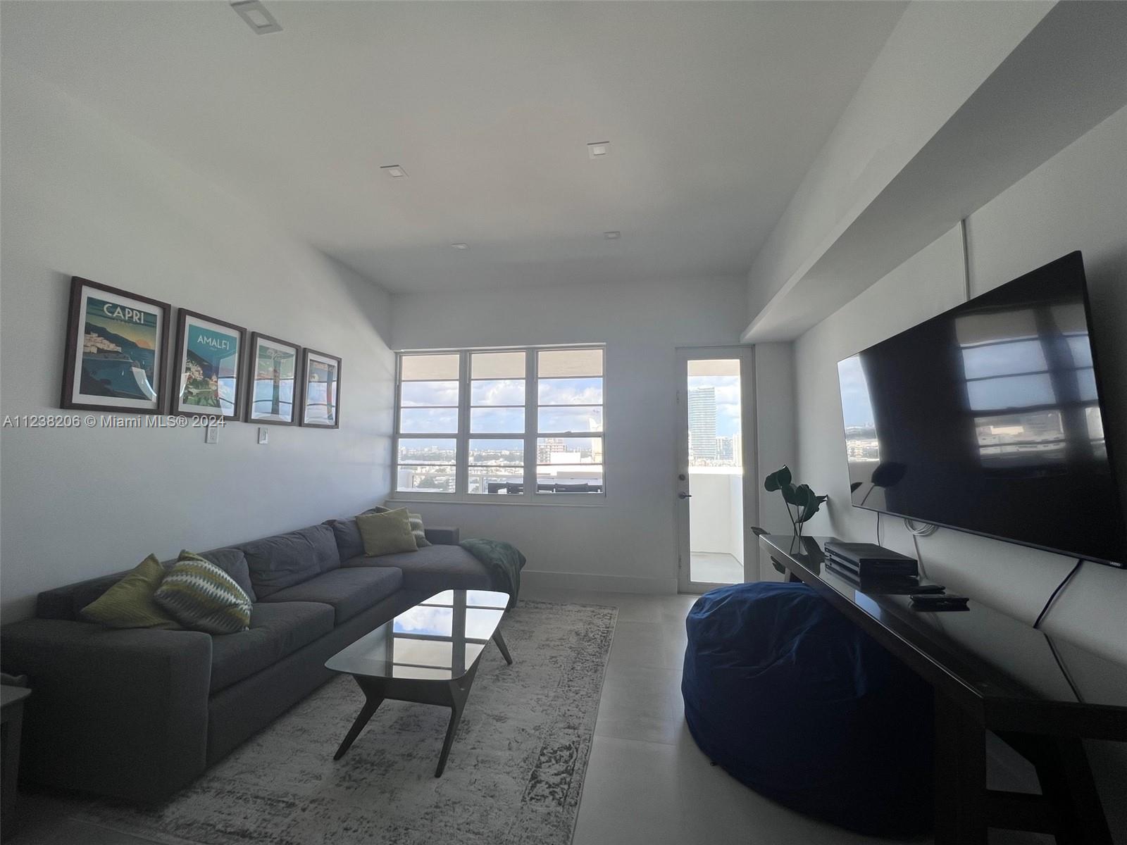 A living room features a grey sectional sofa, a glass coffee table, and a wall-mounted television — 100 Lincoln Rd # 1608, Miami Beach FL 33139, MIAMI BEACH, FL