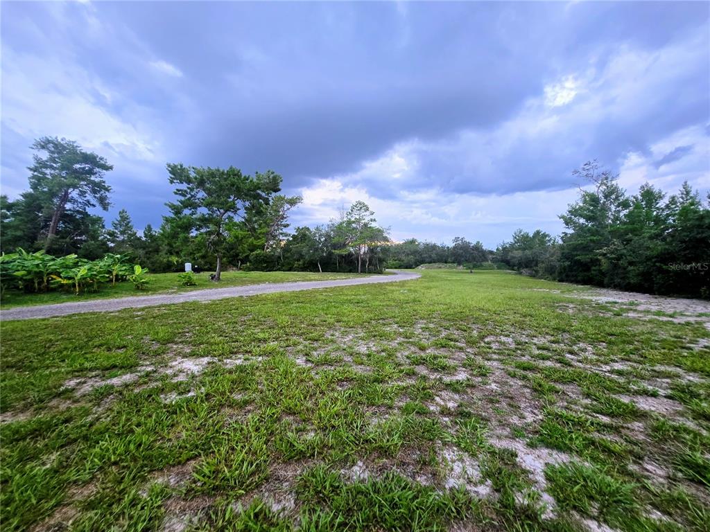 A grassy, partially cleared lot features a gravel driveway and surrounding trees under a cloudy sky — 3130 LORI COURT, DELTONA, FL