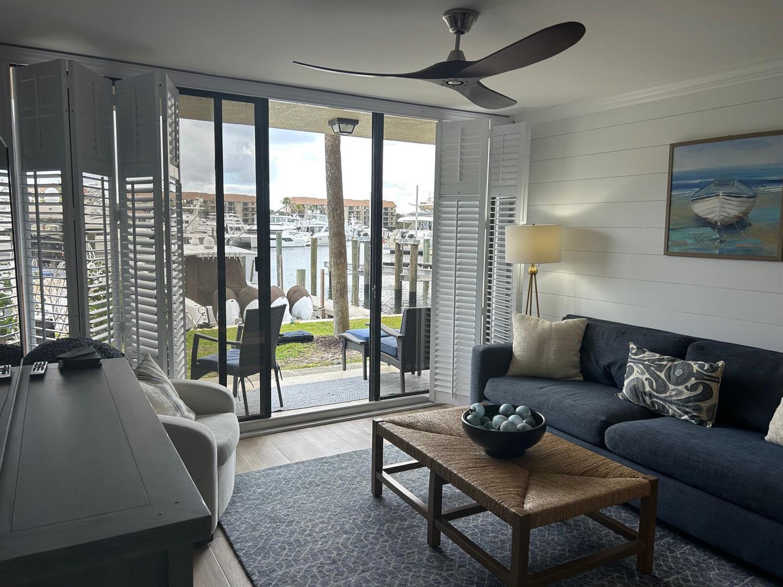 A living room with a dark sofa, woven coffee table, and sliding doors overlooking a marina — 2601 Marina Isle Way # 105, Jupiter FL 33477, JUPITER, FL