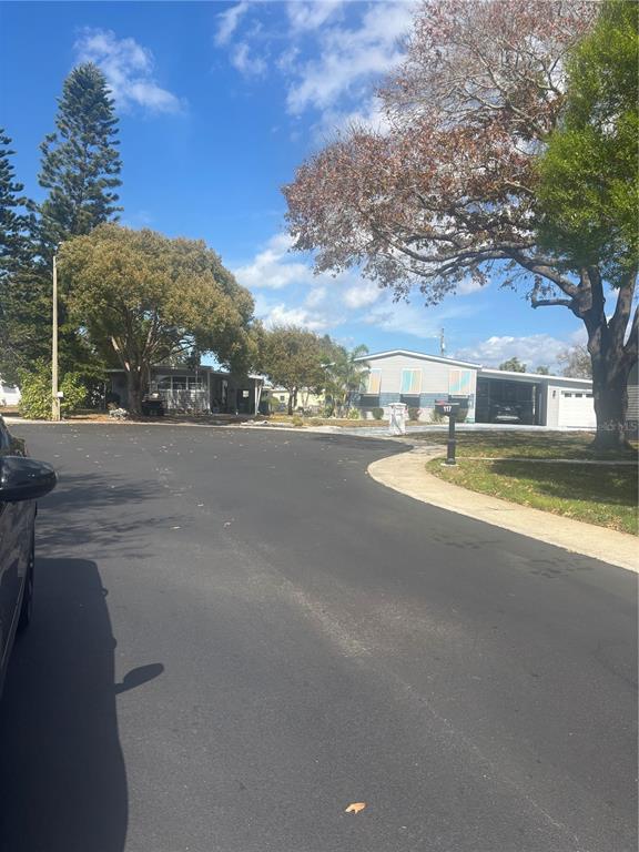A paved street curves past a residential property with a light-colored house and mature trees — 105 FREEDOM COURT UNIT 30, PALM HARBOR, FL