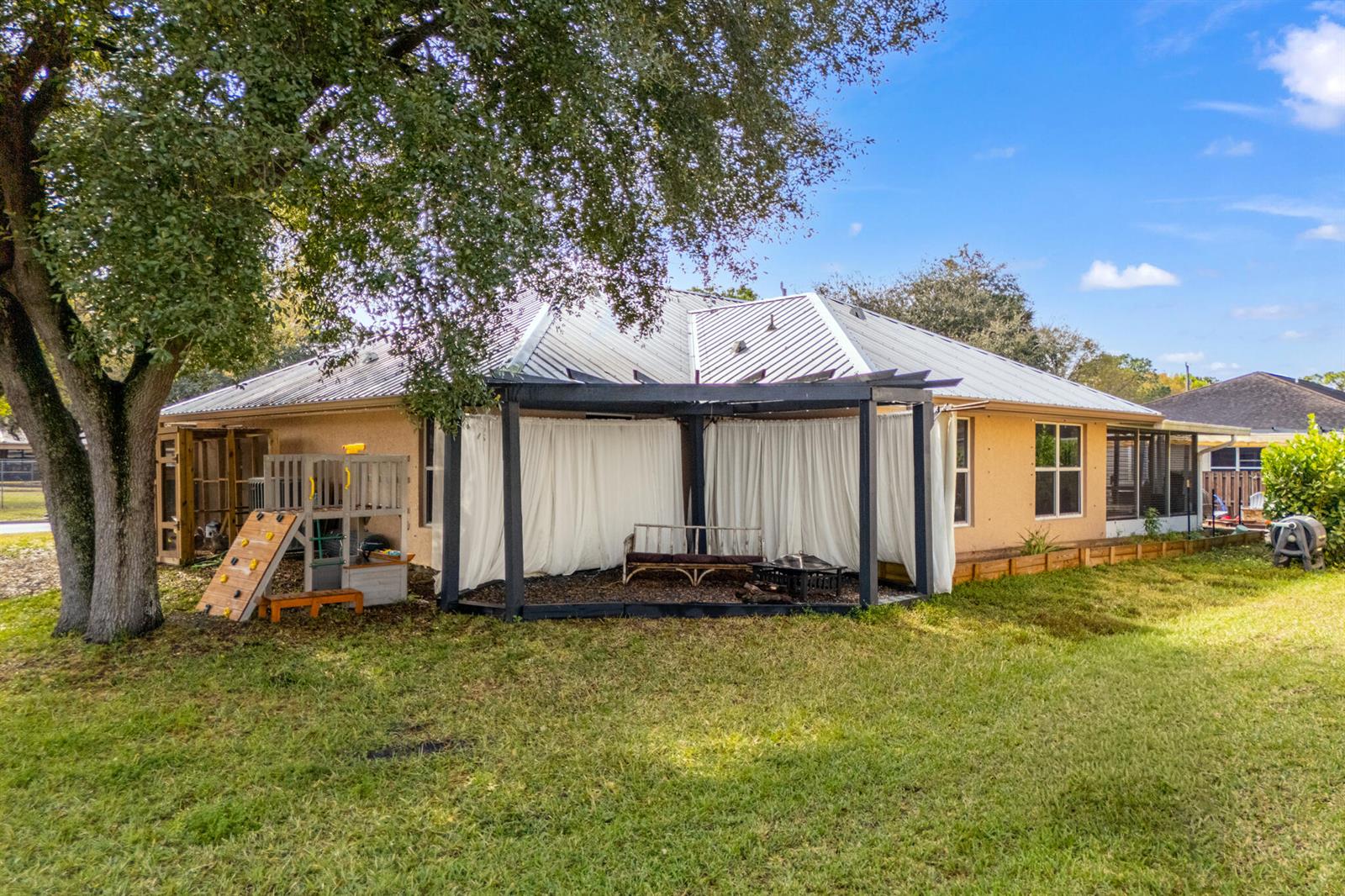 The exterior of a house features a metal roof, a covered patio with curtains, and a playset — 3789 SE 6th Street, Okeechobee FL 34974, OKEECHOBEE, FL