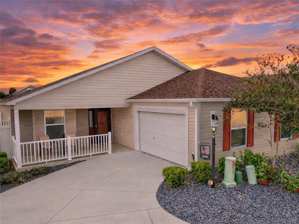A single-story house with beige siding, a white front porch, and a concrete driveway at sunset — 2604 ALCADE PLACE, THE VILLAGES, FL